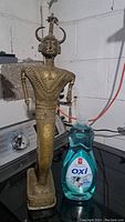 Front view of an old brass statue placed beside a bottle of dish soap for scale, showing detailed geometric patterns on torso and legs, horned headpiece with vertical crest.