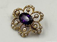 Top view of the brooch on white background showing floral filigree design, central amethyst, and seed pearls