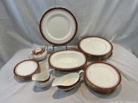 Full view of the Aynsley bone china set arranged showing plates stacked by size, serving pieces including gravy boat, creamer, and lidded sugar bowl. The red and gold border design is clearly visible.