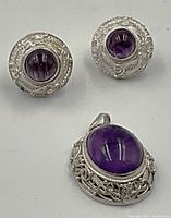 Close up of the silver and amethyst stud earrings and oval pendant showcasing the detailed filigree work and polished purple stones.