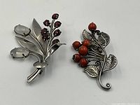 Both sterling silver brooches shown flat on a white surface, one with red faceted gemstones and silver leaves, the other with red beads and silver leaves.