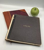 Photo showing two antique map books side by side with a green apple for size reference. One book has a black cover with gold lettering 'ATLAS', the other has a brown leather and maroon cloth cover with gold spine text.