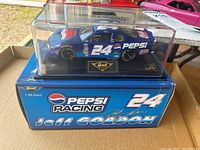 Model car inside clear display case sitting on blue box with Pepsi Racing and Jeff Gordon graphics. The car is blue with white #24 and Pepsi logos.