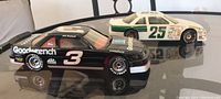 Two NASCAR 1:24 scale die-cast cars on a glass table, front left view showing both Ken Shrader #25 white/green and Dale Earnhardt #3 black cars