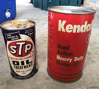 Two vintage sealed oil cans: one Kendall Dual Action Heavy Duty Motor Oil (red) and one STP Oil Treatment (blue/white) on concrete floor.