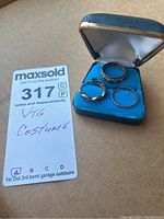 Three metal rings with decorative details displayed in an open vintage blue jewelry case on a brown surface, alongside the lot identification card.
