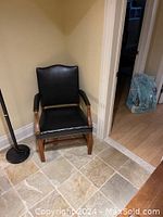 Photo of a black leather armchair with wooden frame and armrests, shown from the front in a corner against a yellow wall and tiled floor.