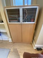 Office credenza cabinet made of beech wood with upper glass doors featuring frosted horizontal stripes. Lower section has two solid wood doors. Cabinet shown standing upright on light wood flooring in a room with white walls.