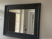 Frontal image of a large black framed rectangular mirror hanging on a light-colored wall, showing the reflection of a door and window frames.
