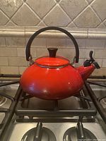 Front and side view of red Le Creuset kettle showing round body, black handle and whistle spout on stove.