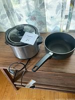Photo of the slow cooker with glass lid and a small black non-stick frying pan on a wooden surface near a window with curtains.