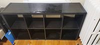 Full view of large black bookshelf with 8 square compartments arranged in two rows of four, placed on wooden floor with visible scuff marks on top surface.