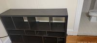 Black wooden book shelf against white wall and near bathroom entrance. Multiple cubby compartments on top and two lower storage cabinets with doors open and closed.