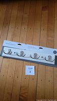 Photo showing brand new packaged white wooden coat rack with four double metal hooks, placed on a wood floor.