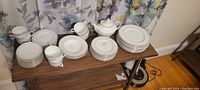 Photo of the entire formal dinner set arranged on a wooden surface, showing stacks of plates, bowls, tea cups with saucers, and a teapot. The pieces are white with a subtle decorative trim.