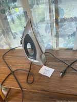Photo of one grey and black electric steam iron with power cord placed on a wooden surface, near a floral patterned curtain.