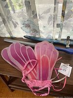 Photo showing two pink kids fairy wings with elastic straps and two toy swords, placed on wooden furniture near window with patterned curtains.