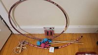 Photo of a kids' games lot including a pink and black patterned hula hoop, matching toy bow with blue and pink soft tip arrows, and a natural fiber jump rope with black handles, all laid out on a wooden floor.