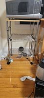 Front and slight angle view of a metal kitchen cart with wood top, three metal wire shelves, and caster wheels. The cart is standing on a wooden floor with a microwave placed on top.