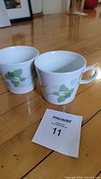 Photo showing two white ceramic mugs with green leaf pattern on a wood floor with a MaxSold tag with number 11.