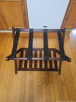 Photo of a wooden luggage rack with three black fabric straps on top surface, and a wooden slatted shoe shelf below. Positioned on a wooden floor, against a white wall and wooden cabinets.