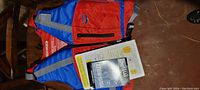 Youth life jacket front view showing blue and red color scheme, front zipper pocket, and attached safety information booklet.