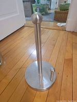 Paper towel holder made of stainless steel, placed on wooden floor showing base and vertical rod with ball finial top.