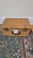 Closed wicker picnic basket with leather straps and handle on a patterned rug.