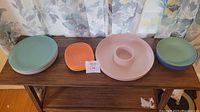 Photo showing plastic dishware items arranged on a wooden table: 5 pastel light green plates stacked on left, 1 orange square dish in center-left, large round light pink and white divided dip dish in center, 4 green bowls stacked on right.