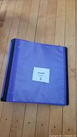 Purple plastic school binder with black spine laying closed on wooden floor.