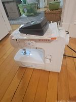 White Singer sewing machine on hardwood floor with foot pedal and manual resting on top.