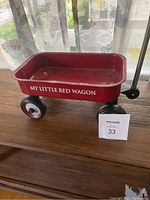 Photo shows the small red wagon positioned on a wooden surface near a window, showcasing the side with the text 'My Little Red Wagon'. The wagon has four black and white wheels and a black handle extending to the side.