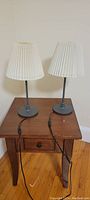 Two IKEA table lamps on a wooden table showing white pleated shades, black stands, and gray round bases with cords