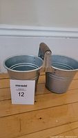 Galvanized metal double utensil holder with wooden handle placed on wooden floor against white baseboard.