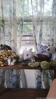 Photo of various stuffed animals placed on a wooden surface near a window with floral curtains in the background.