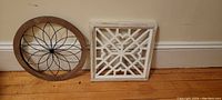 Two pieces of wall decor shown leaning against a wall on floor, one round with wooden frame and metal design, one square with white geometric lattice frame.