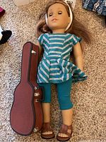 Front view of American Girl doll in striped top and teal leggings lying next to brown guitar case.