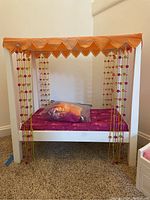 Front view of the American Girl doll bed showing white wooden frame, pink cushion, beaded curtains, and packed pillow on bed.