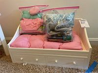 White wooden American Girl doll day bed with pink mattress pad and bags containing extra bedding and books on top