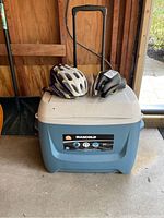 Three bike seats, one helmet on top of Maxcold cooler with wheels and pull handle.