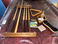 Five wooden billiard cues with cue rack, triangle rack, brush, chalk, and billiard books laid out on table.