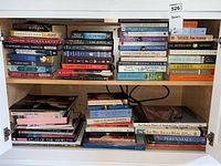 Books stacked on a wooden shelf, mostly novels and some coffee table books visible