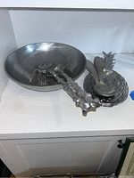 Front view of metal salad bowl with metal serving tongs and pineapple-shaped serving platter with utensils