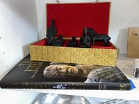 Five black Chinese Terracotta Soldier figurines including a horse displayed in a gold and red velvet lined box placed on top of a book titled 'The Eternal Army'.