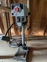 Front view of Delta Shopmaster drill press showing the machine head with speed settings, feed handles, power switch, and base with adjustable table and lamp.