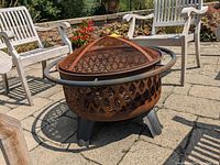 Angled side view of a rusty metal fire pit with a circular mesh cover and black ring handle.