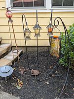 Outdoor garden setting with three black metal shepherd hooks each holding a lantern and a curved metal stand with a stained glass hanging lantern. Also visible are a metal garden stake with a glass ball top and two small metal animal figurines on the ground.