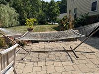Front and side view of the LLBean metal base hammock stand with attached white/beige net hammock and cushion. Shows stand on stone patio with plants and lawn in background.