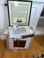 Framed cross stitch art with poem and packaged jewelry box with shower curtain liner on top, against a white wall and white baseboard.