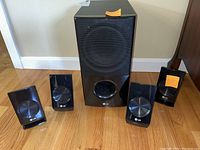 Wide shot showing the large subwoofer speaker and the four smaller satellite speakers placed on the floor against a wall.